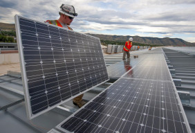 Two men are standing on a roof installing solar panels, with a beautiful sky and mountains in the background.