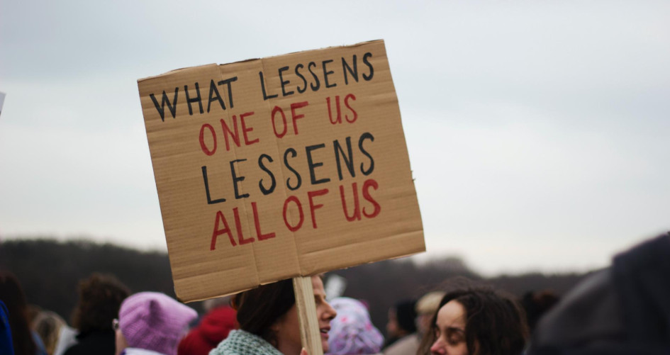 Image of a woman holding a sign that reads "What lessens one of us lessens all of us"