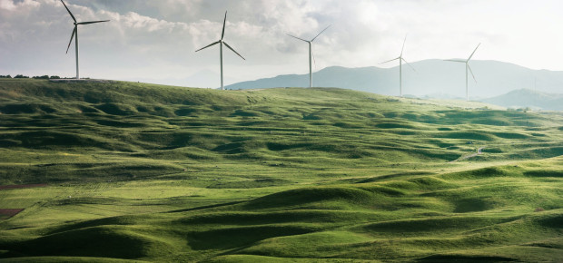 A row of windmills stand atop a bank of grassy, green hills.
